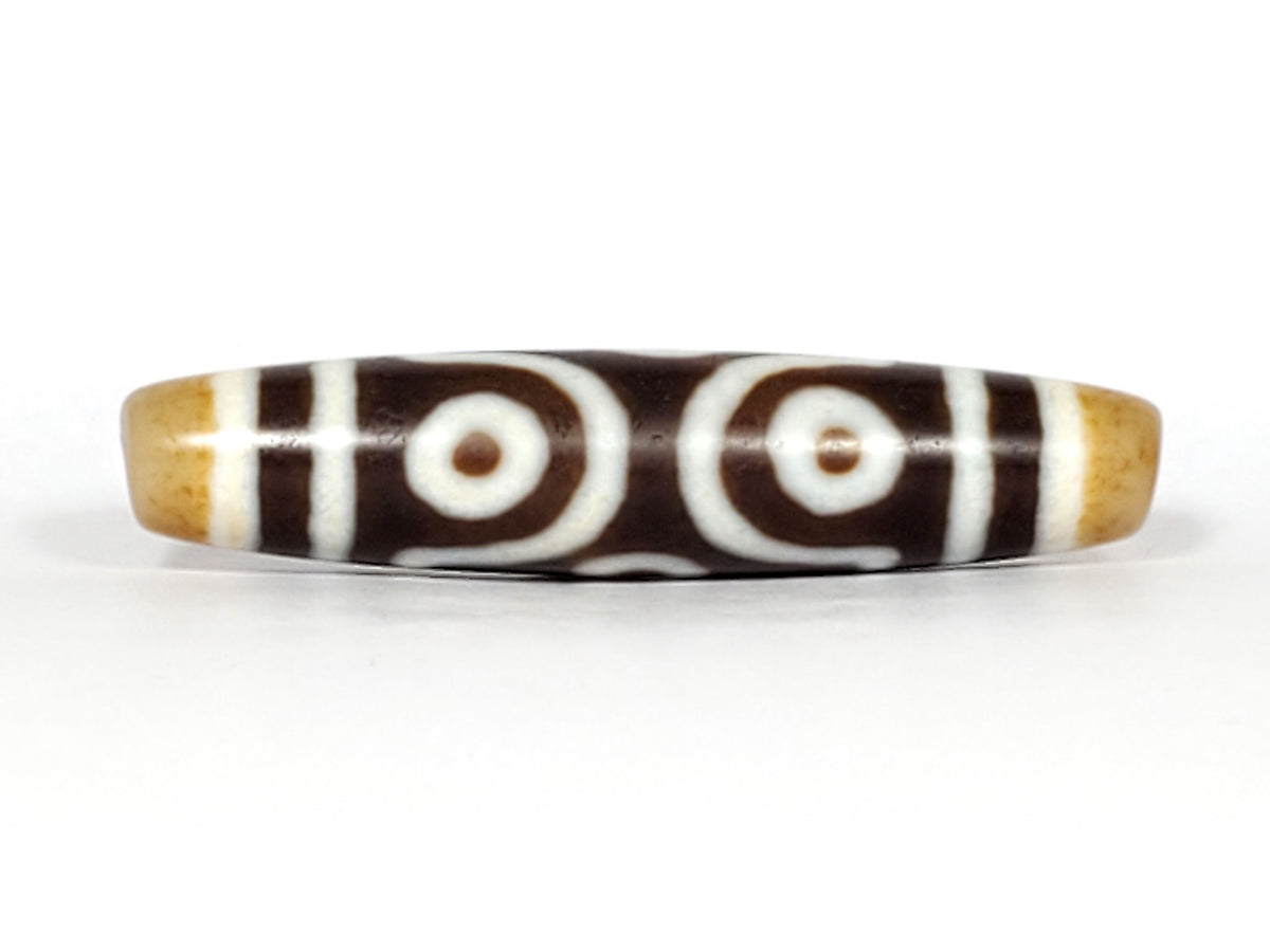 Three-quarter view of antique six eye dzi At-053026-6E, honey-gold end cap with drill hole, two closed eyes visible, elongated torpedo body, deep brown agate, 54.9mm, unretouched