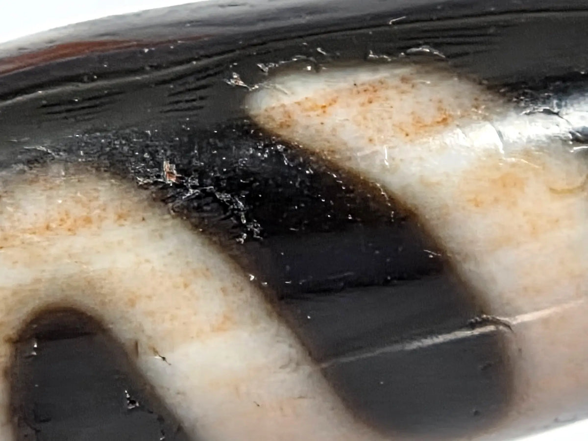 Macro close-up of antique Tibetan dzi bead AT-B-101226-MH showing surface flaking and spalling damage in dark agate zones — thin surface layers partially lifting and fragmenting, rough fractured texture visible alongside warm amber bloodspot staining in cream zones