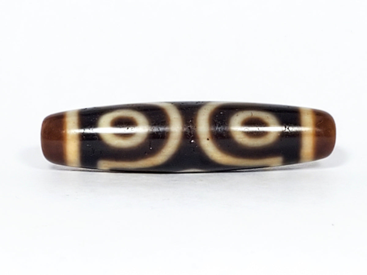 Antique Tibetan three-eye dzi bead, dark agate with cream eye motifs, brown patina, elongated barrel shape, aged surface texture
