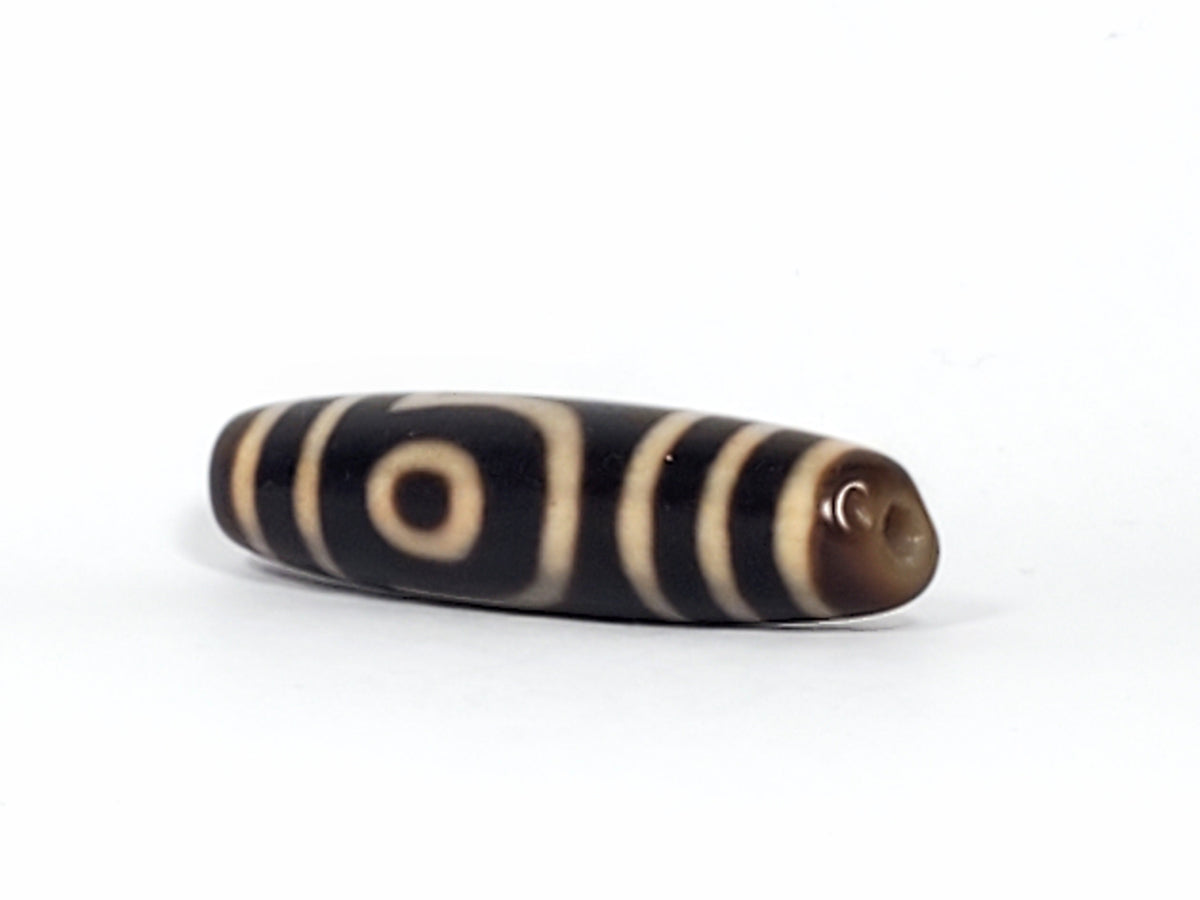 Antique two-eye dzi bead, dark agate, cream stripe and eye motifs, visible drill hole, natural patina, elongated barrel shape