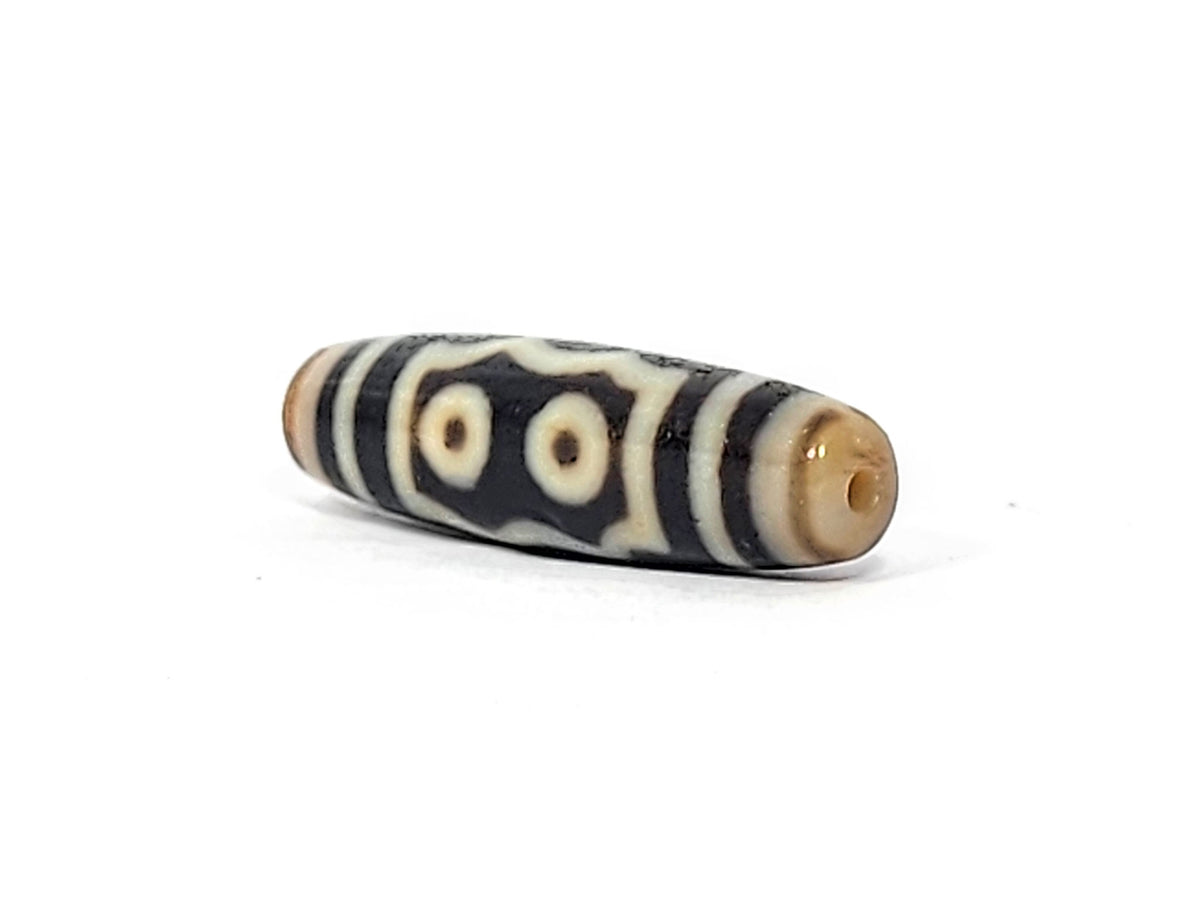 Three-quarter view of antique five eye dzi At-072625-5E, warm honey-tan end cap with drill hole, two-eye frame composition visible, pitch-black torpedo body, unretouched