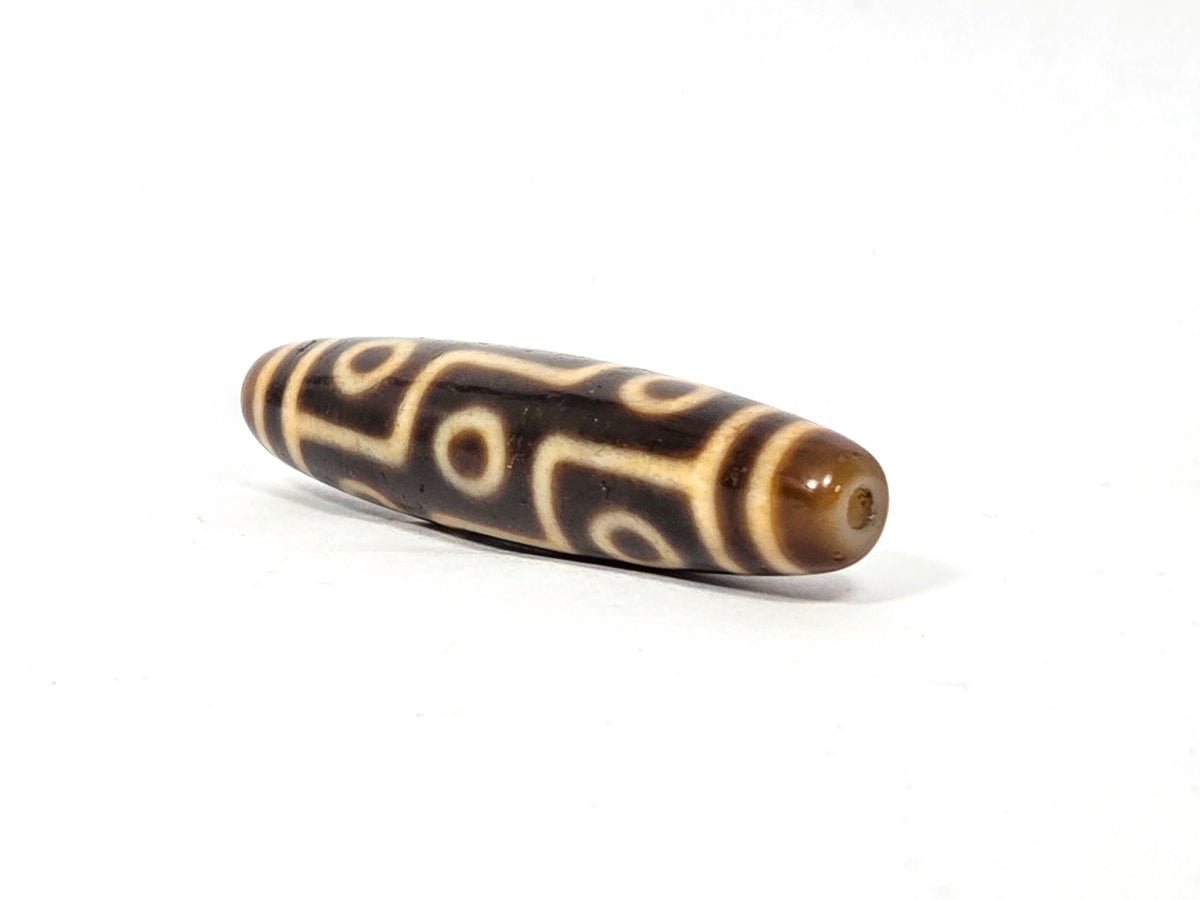 Antique Tibetan nine-eye dzi bead, dark brown agate, cream eye motifs, aged patina, visible drill hole, elongated barrel shape