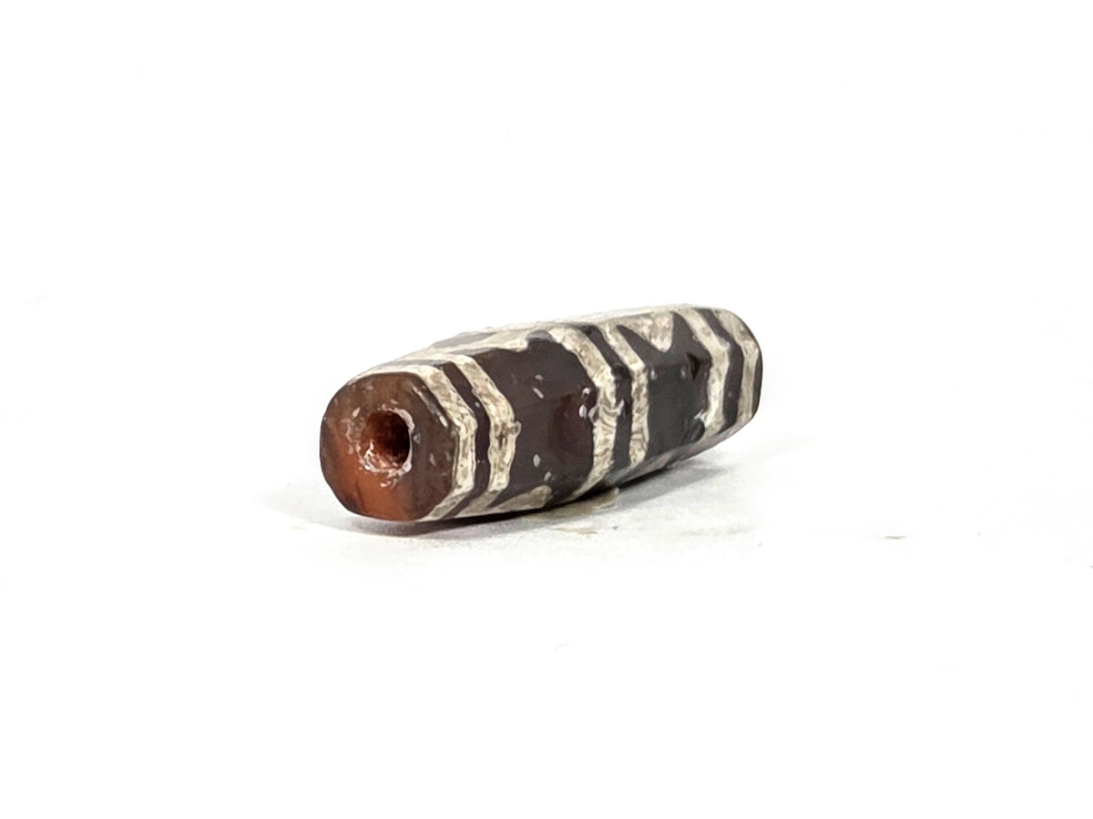 Opposite angled end view of ancient etched carnelian bead showing second drill hole in warm carnelian tip, full octagonal end face visible, crystalline white etching bands across faceted body, 700–1000 years old