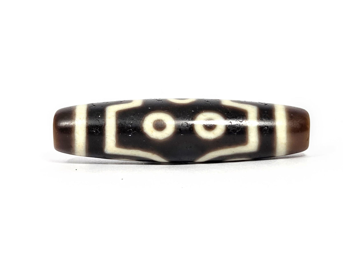 At-072226-7E Seven Eye face B — two central eyes in geometric frame, pitch-black ground, dark brown end caps, unretouched.