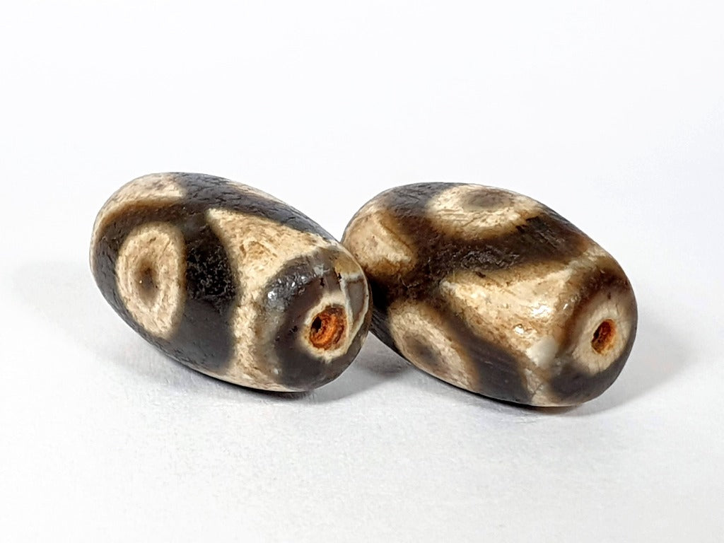 "Pair of New Faux Antique Three Eye Motif Dzi Beads" Description: "A pair of new faux antique Dzi beads with a three eye motif. These beads emulate the look of ancient Dzi beads, offering a stylish and symbolic accessory."