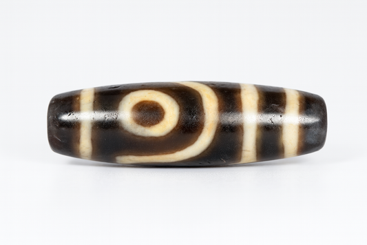 Ancient Tibetan Dzi bead with two eye motifs, featuring a dark brown base and cream-colored circular patterns, centered against a white background.