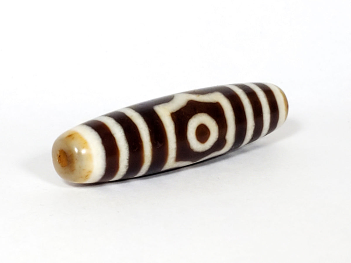 Three-quarter view of antique three eye dzi At-053126-3E, warm honey-tan end cap with drill hole, octagonal-frame eye visible, chocolate-brown torpedo body, unretouched