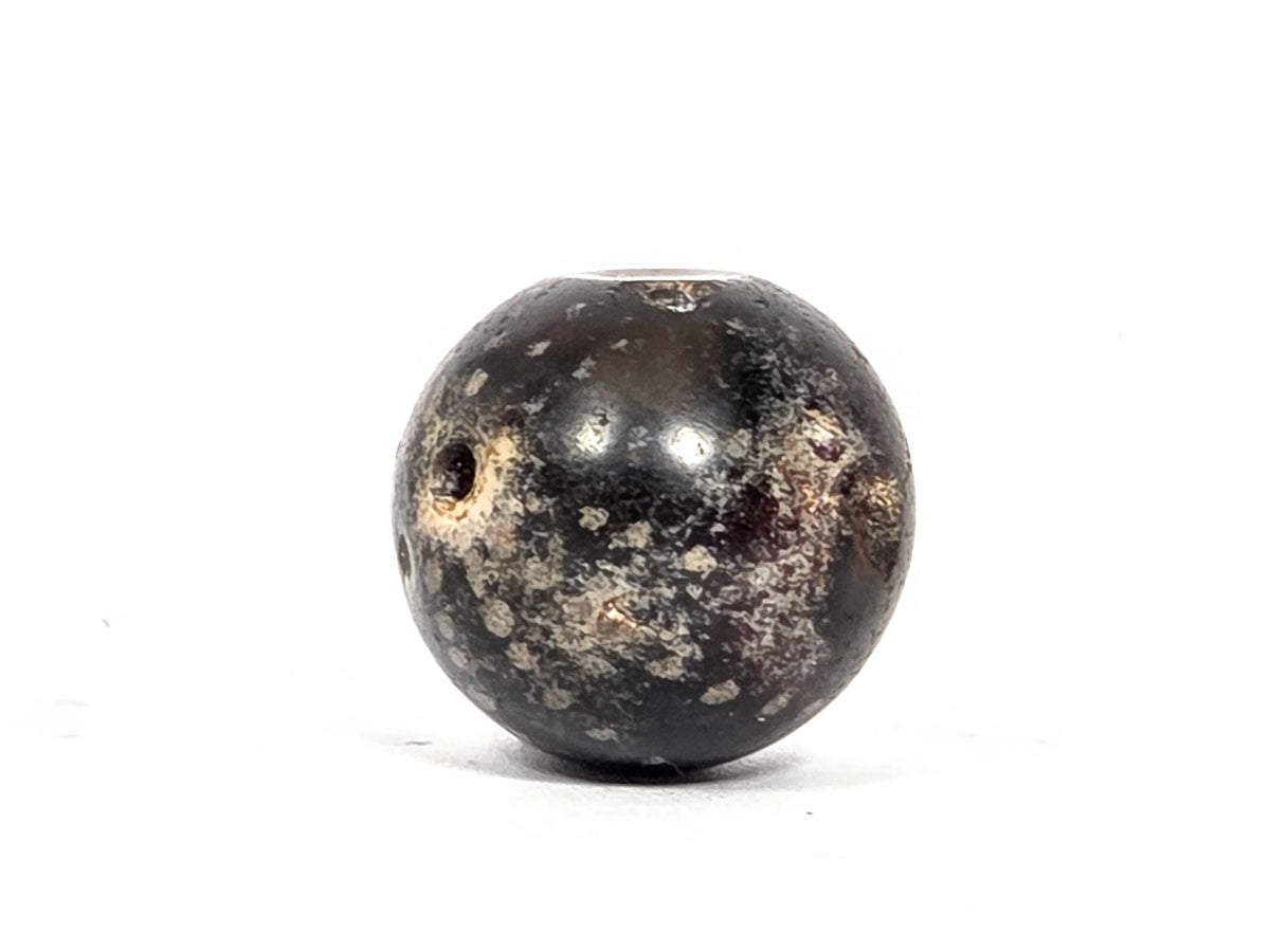 Opposite end view of ancient round agate bead AC-101126-RS showing second drill hole in deep charcoal-grey hemisphere with extensive cream-grey calcification spreading broadly around the aperture, 1500–2000 years old
