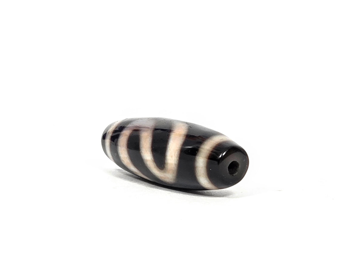 Angled end view of antique Tibetan dzi bead AT-B-101226-MH showing drill hole aperture at pitch-black tip with cream band end cap and diagonal money hook motif lines on black agate body