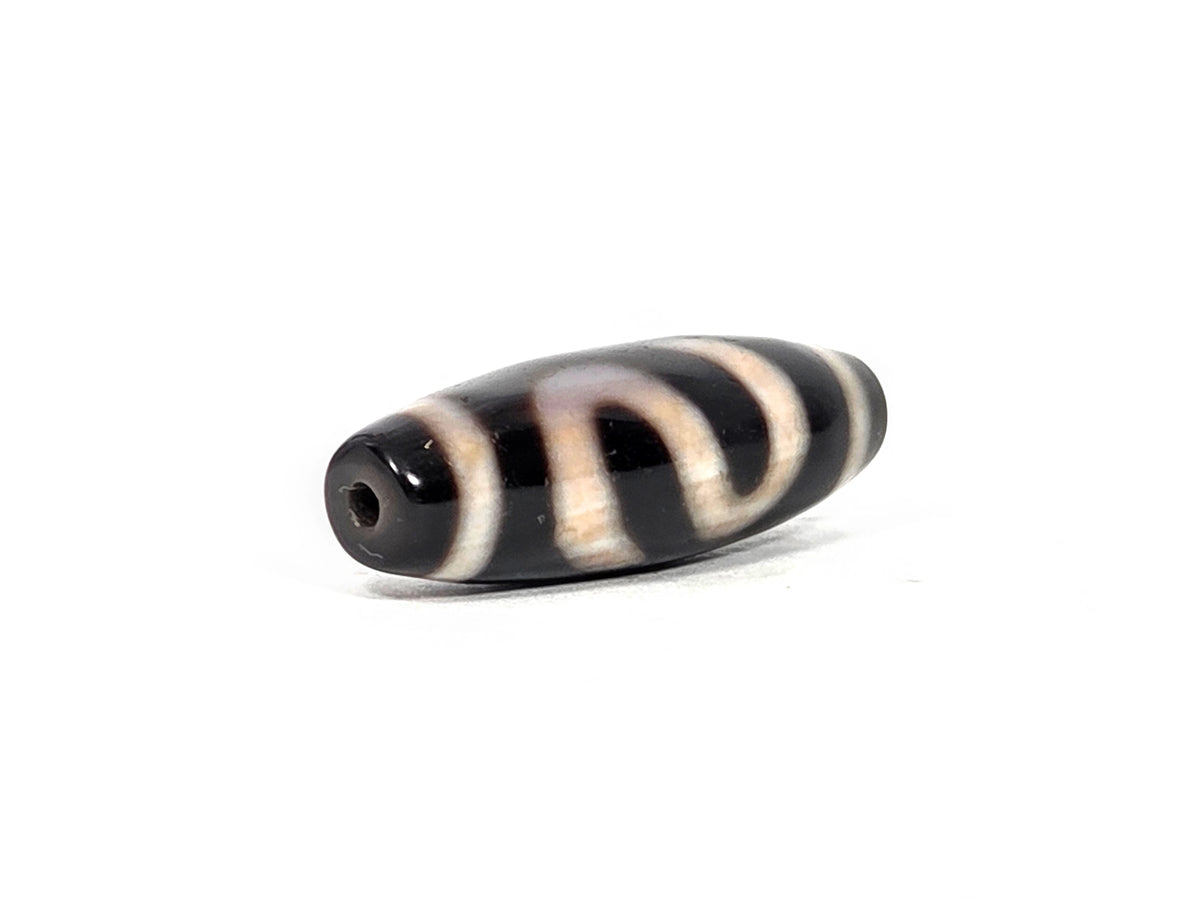 Opposite angled end view of antique money hook dzi bead showing second drill hole at pitch-black tip, full barrel profile, and cream band end cap with money hook motif lines on deep black agate, 100–300 years old