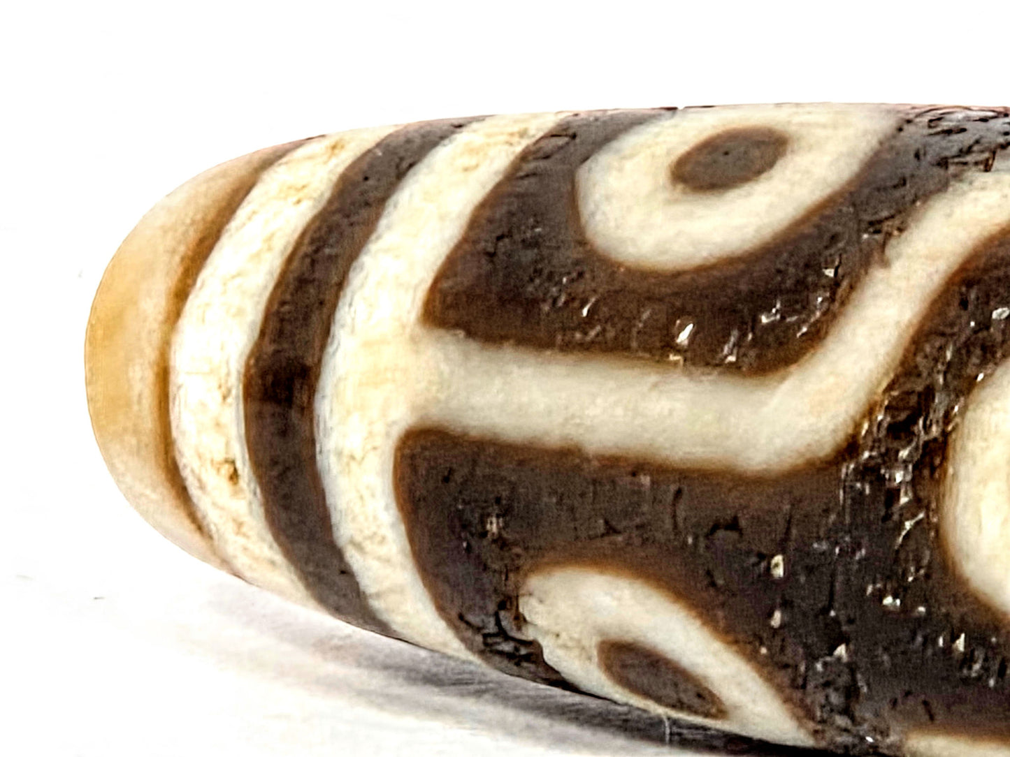 Close-up of ancient Nine Eye Dzi bead surface — warm tan-amber white agate texture and deep brown etching detail visible, ~800 years of surface character