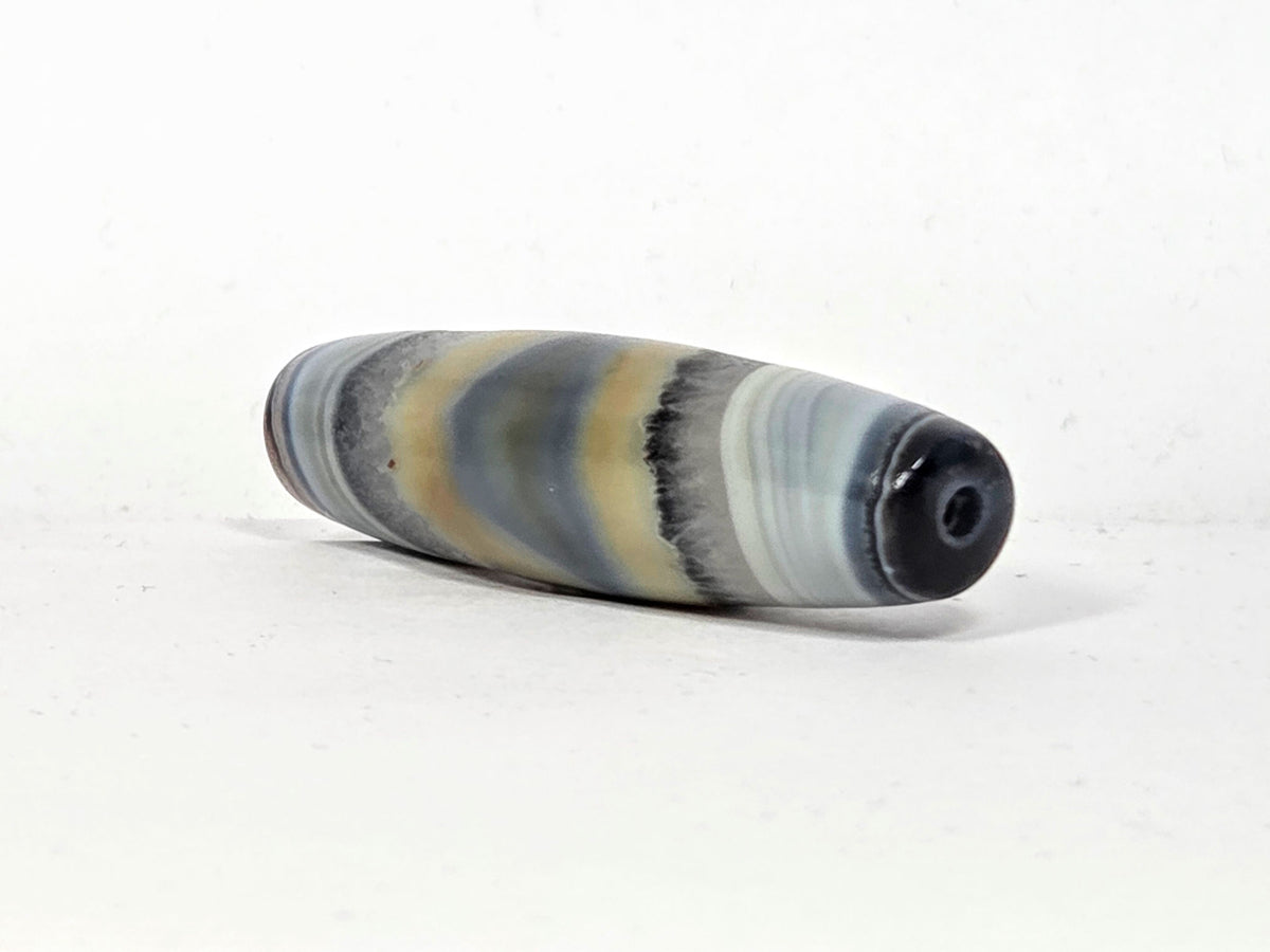 Antique one-eye Dzi bead, elongated barrel shape, grey-blue banded agate, natural patina, visible drill hole, tan stripe motif