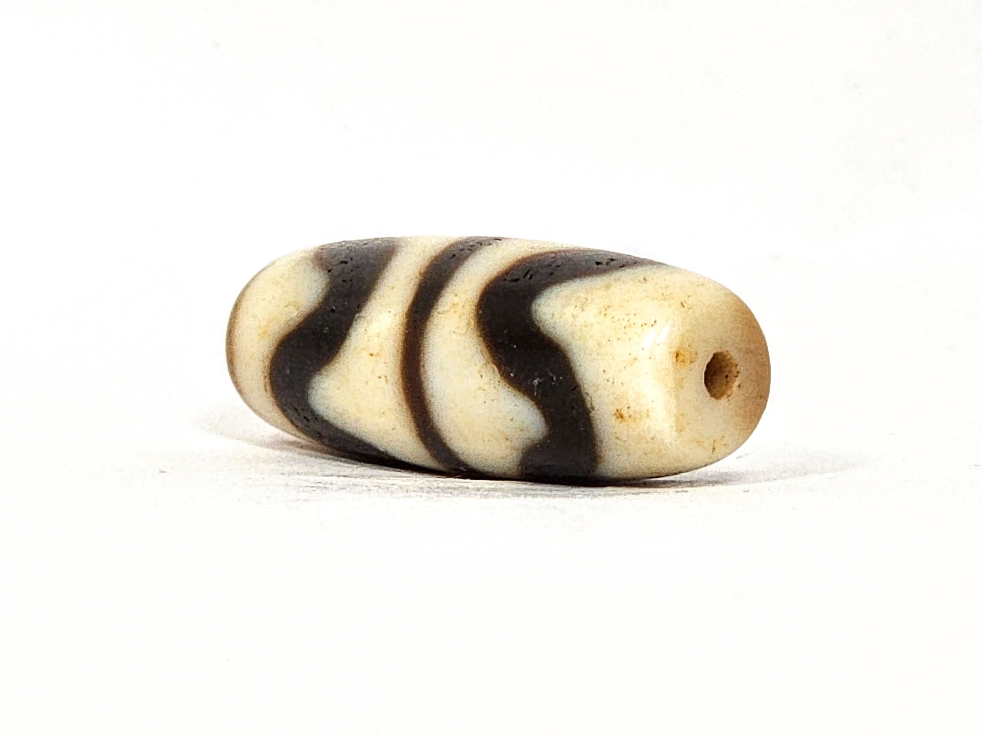 Three-quarter view of antique Double Wave dzi At-072325-DW5, warm brown end cap with drill hole, wave motif visible on compact torpedo body, 28.4mm × 10.6mm, unretouched
