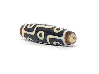 Antique Tibetan nine-eye dzi bead, dark agate with cream white eye motifs, visible drill hole, aged patina, elongated barrel shape