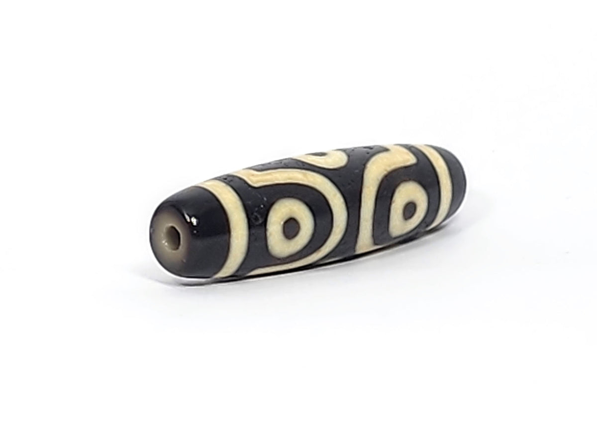 Three-quarter view of antique six eye dzi At-042925-6E2, pitch-black end cap with drill hole, two C-ring eyes and scroll visible, elongated torpedo body, unretouched
