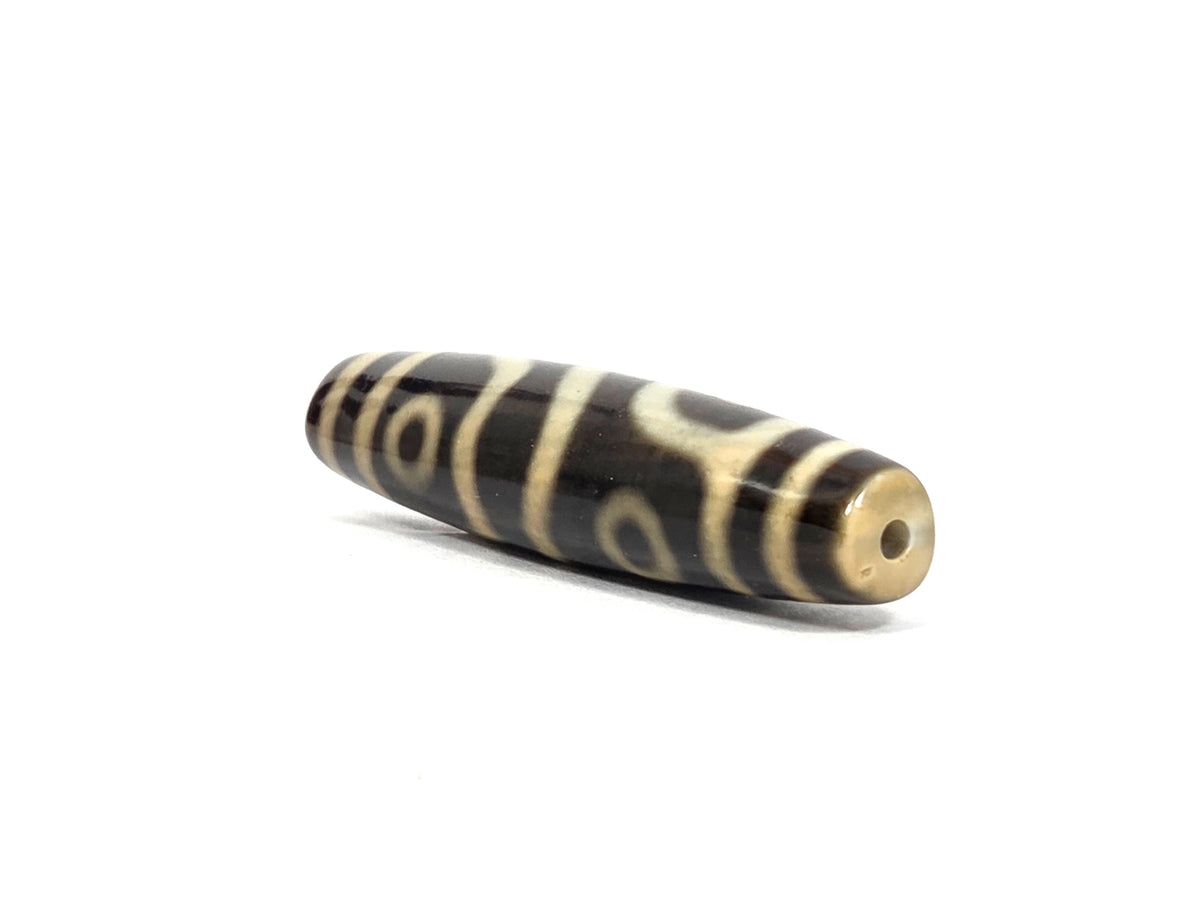 Angled end view of new four eye variant dzi bead N-102526-4EVS showing a centered drill hole in a translucent honey-amber agate tip, with framed cream eyes and curving motif lines along the dark brown barrel
