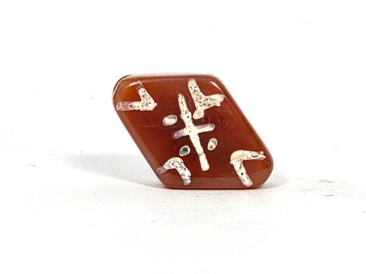 Reverse diamond face of ancient carnelian cross and tiger tooth motif bead showing full composition: cross center, corner bracket elements, and cream-white etched lines on deep amber-red carnelian ground, 500–700 years old