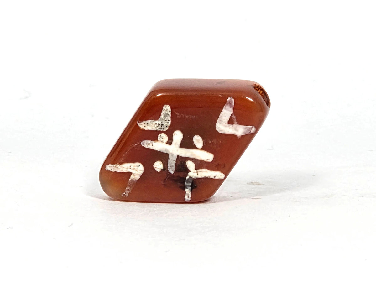 Ancient etched carnelian bead AC-101026-CM3, diamond-face view showing central cross motif with four corner tiger tooth brackets and dot accents on deep amber carnelian ground, 500–700 years old