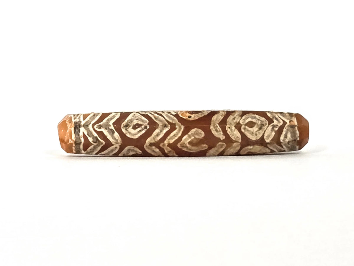 Ancient etched carnelian bead AC-101426-12E, first lateral profile showing dense cream-white twelve-eye and tiger wave motif — diamond-framed eye units alternating with chevron zigzag band lines across warm reddish-orange carnelian ground, eight-sided faceted cylinder form, 1500–2000 years old