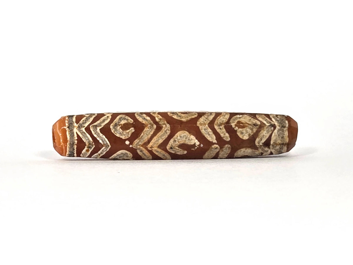Second lateral profile of ancient etched carnelian bead AC-101426-12E showing reverse face with dense cream-white tiger wave chevron bands and diamond-framed eye units covering warm reddish-orange carnelian ground, elongated eight-sided faceted cylinder, 45.2 mm × 9.3 mm