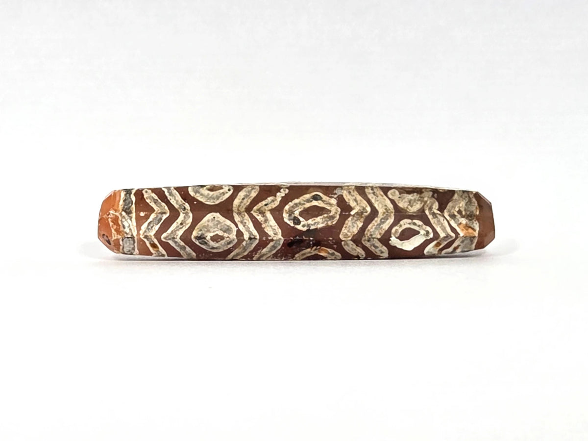 Third lateral profile of ancient etched carnelian bead AC-101426-12E showing octagonal faceted form clearly — flat facet planes and ridges between faces visible at edges, full-surface cream-white twelve-eye and tiger wave etching on warm reddish-orange carnelian, 1500–2000 years old