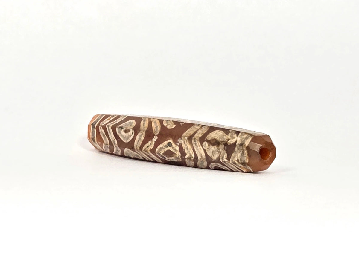 Opposite angled end view of ancient etched carnelian bead AC-101426-12E showing second drill hole at warm orange carnelian tip, octagonal cross-section, and full-surface cream-white tiger wave and twelve-eye etching running close to the tip aperture, 1500–2000 years old