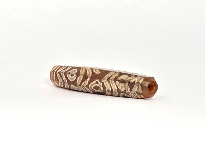 Opposite angled end view of ancient etched carnelian bead AC-101426-12E showing second drill hole at warm orange carnelian tip, octagonal cross-section, and full-surface cream-white tiger wave and twelve-eye etching running close to the tip aperture, 1500–2000 years old