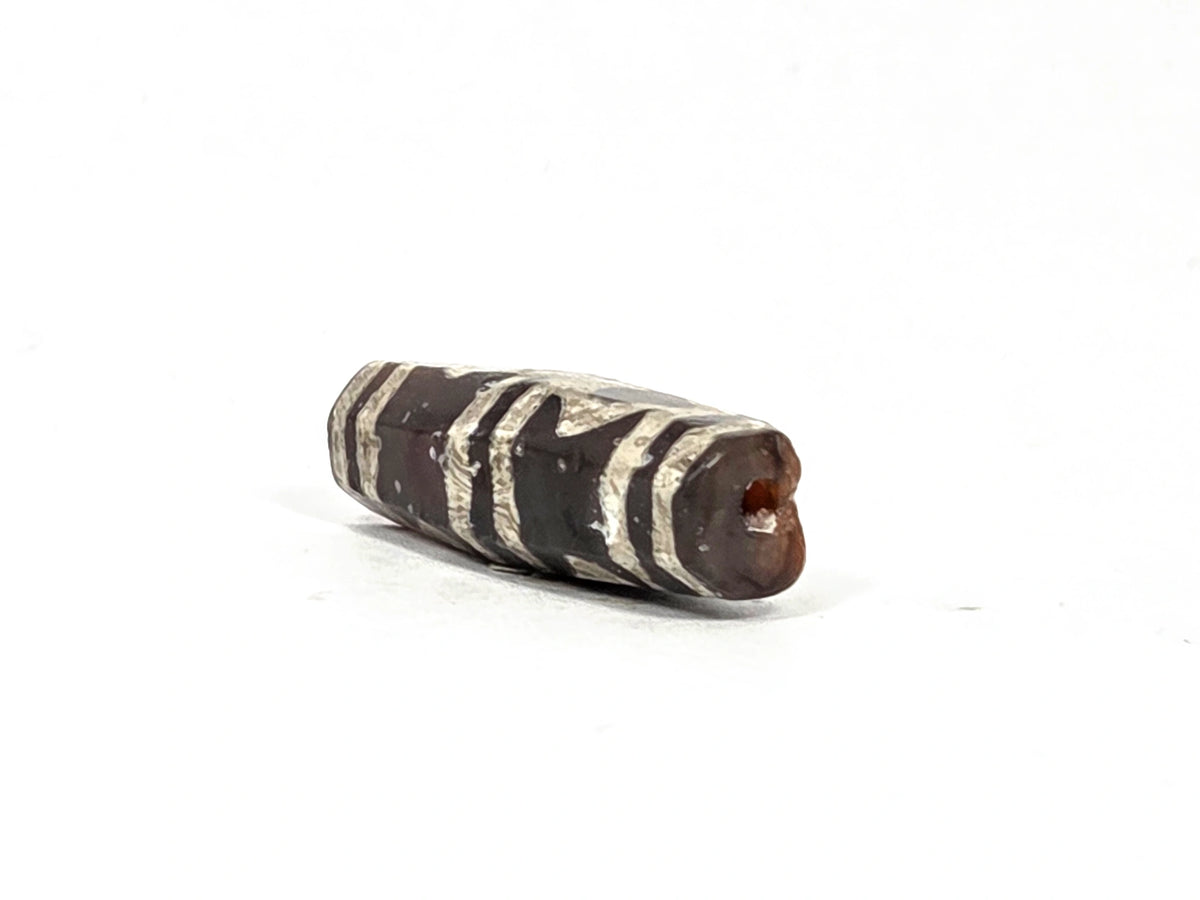 Angled end view of ancient etched carnelian bead AC-101226-EC showing drill hole aperture in warm reddish-orange carnelian tip, octagonal cross-section visible, silver-white etching bands along faceted barrel body