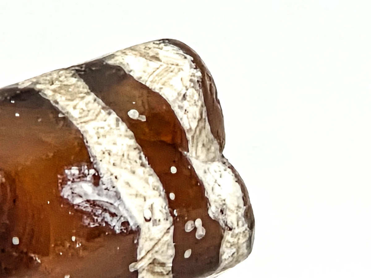 Extreme macro close-up of ancient etched carnelian bead AC-101226-EC showing granular crystalline texture of silver-white lime etching against warm translucent amber-brown carnelian ground, 700–1000 years old