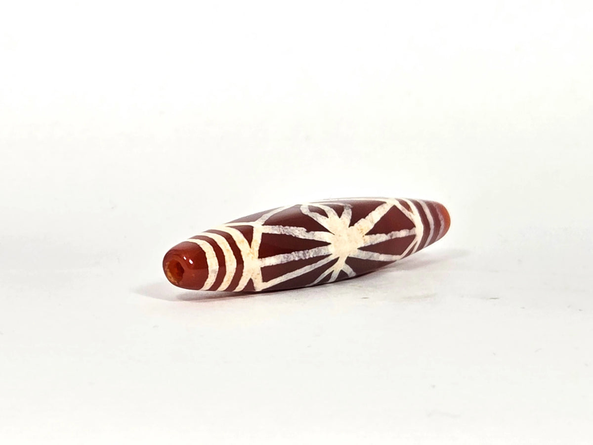 Angled end view of ancient carnelian starburst bead showing drill hole aperture set in warm amber-orange carnelian tip, with band lines and radiating motif visible, 500–700 years old