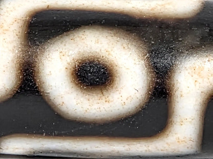 Extreme macro of a single eye on ancient Tibetan nine-eye dzi bead showing concentric cream-white ring, deep black center, and natural bloodspot brown patina at the ring edges