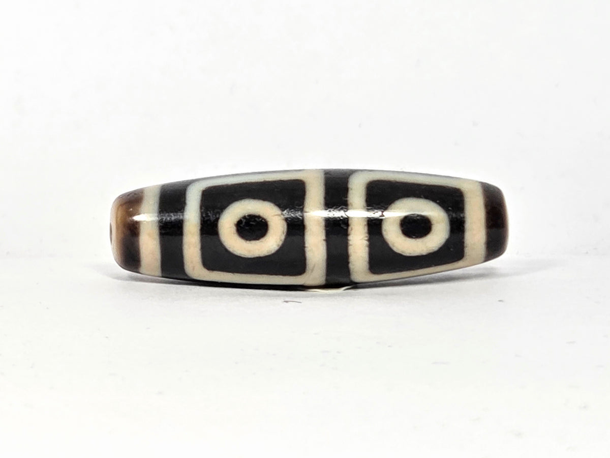 Antique Tibetan dzi bead, four-eye motif, black and cream agate, oval barrel shape, etched circles, natural patina, visible drill hole.