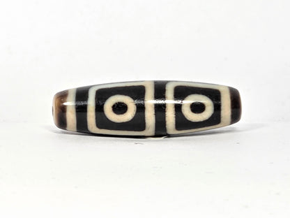 Antique Tibetan dzi bead, four-eye motif, black and cream agate, oval barrel shape, etched circles, natural patina, visible drill hole.
