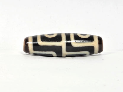 Antique Tibetan dzi bead, four-eye motif, black and cream agate, geometric white lines, oval barrel shape, aged patina visible.