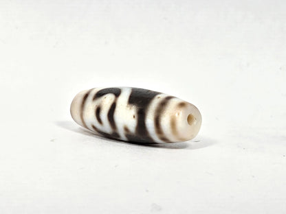 Angled view of antique Tibetan dzi bead AT-B-101226-TLV showing elongated profile with dark horizontal stripe bands, cream-white agate body, and drill hole aperture at cream-white tip, 200–300 years old