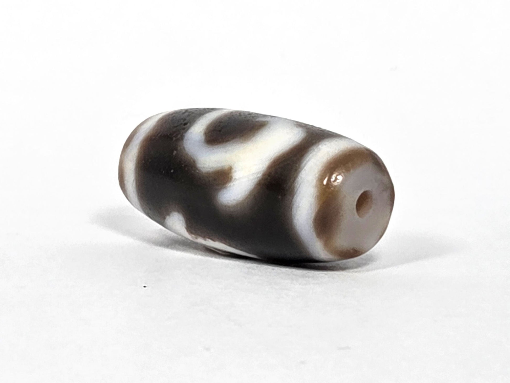 At-080926-RuyiBS antique Ruyi dzi bead 26.2×12.0mm, angled drill-end view showing warm grey-brown end cap, cream shoulder stripe, and body motif, unretouched