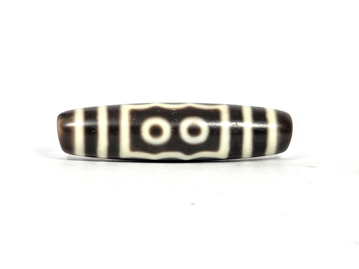 Two-eye face of antique five-eye dzi bead (AT-100826-5E) showing two cream-white eye rings within a rectangular cream border with wavy top and bottom edges on dark brown agate