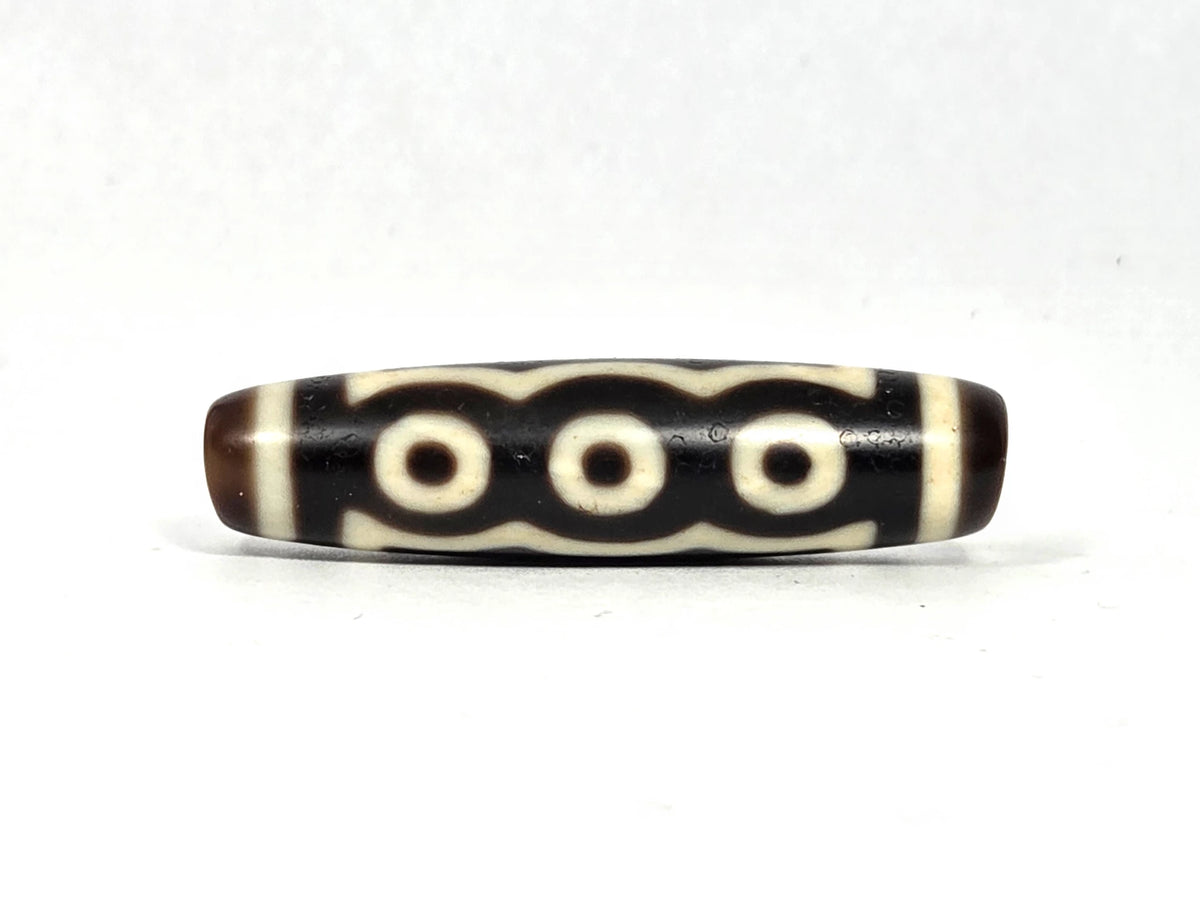Antique Tibetan five-eye dzi bead (AT-100926-5E2), three-eye face showing three cream-white eye rings beneath a scalloped cloud-wave line on deep dark brown and black agate, 48.9 mm, 200–400 years old