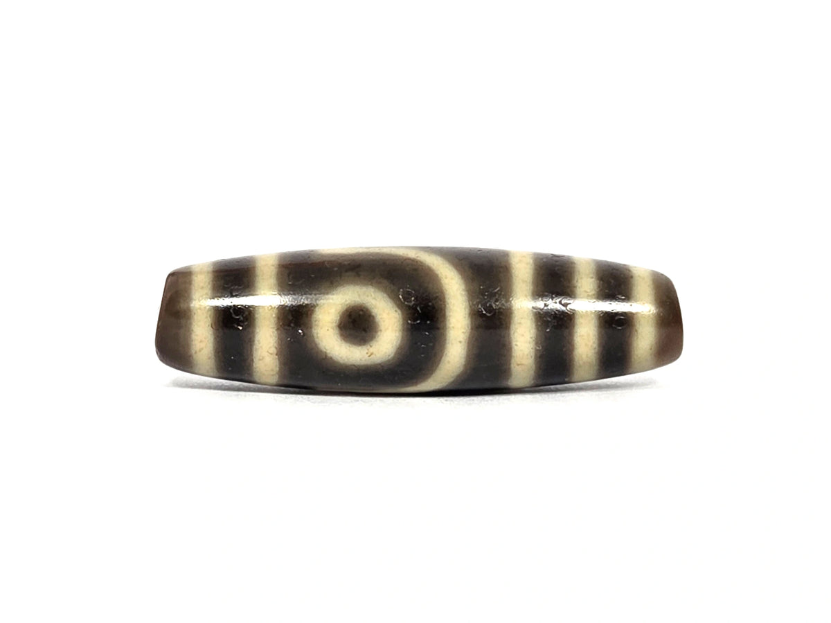 Antique Tibetan two eye dzi bead AT-102126-2E, first face — cream-white eye with dark pupil framed by a curved cream line, cream bands at both ends, on dark brown agate that photographs near-black, 200–400 years old