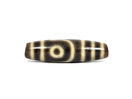 Antique Tibetan two eye dzi bead AT-102126-2E, first face — cream-white eye with dark pupil framed by a curved cream line, cream bands at both ends, on dark brown agate that photographs near-black, 200–400 years old