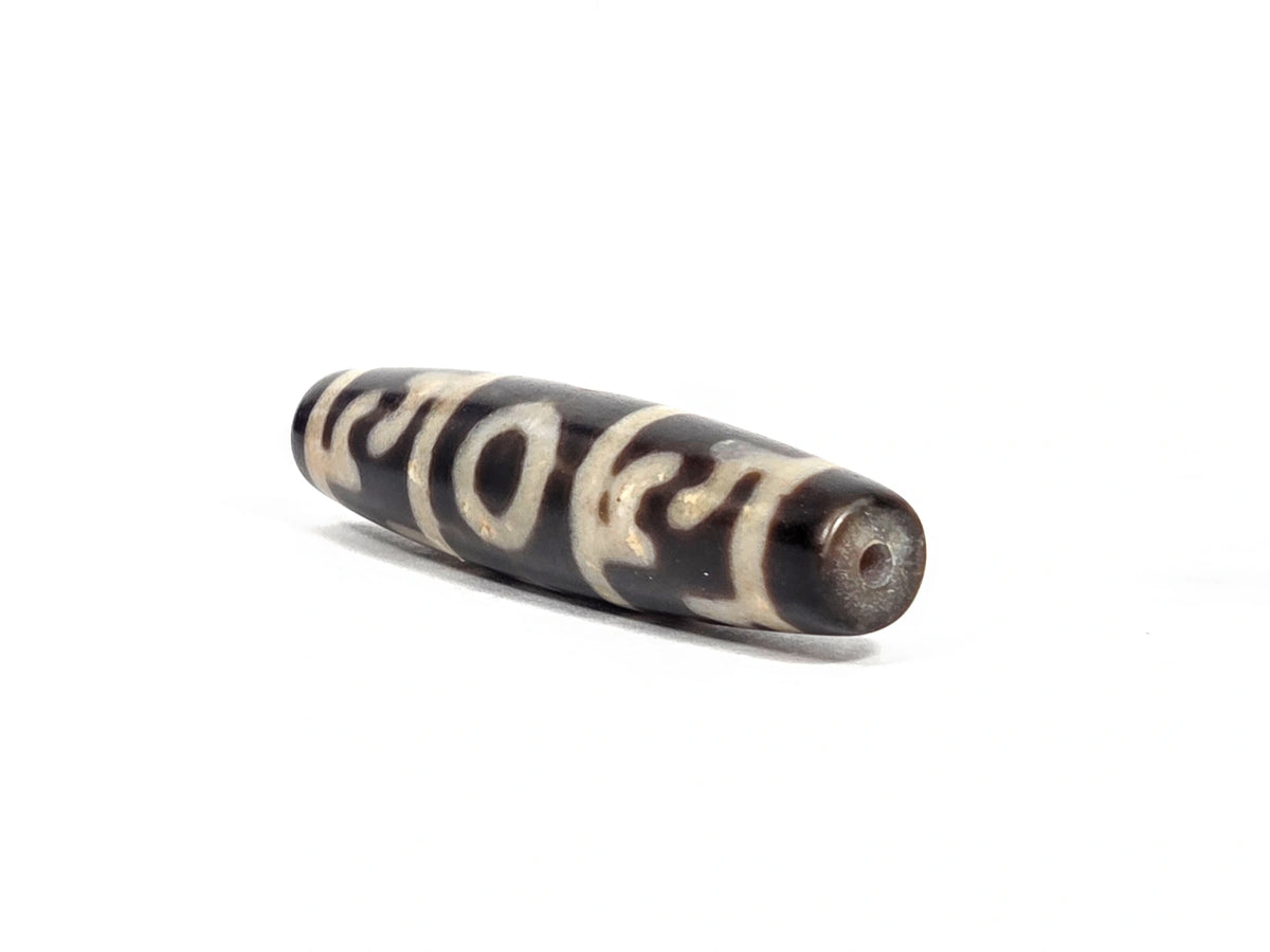 Angled end view of new Taiwan three-eye Guru Rinpoche paraphernalia dzi bead (N-092726-GRP3E) showing small clean drill hole set in deep brown-black agate tip, elongated torpedo form visible