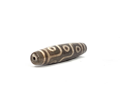 Angled end view of new nine-eye dzi bead showing drill hole aperture in dark grey-brown agate tip with band lines and eye pattern visible along the body, 58.5 mm