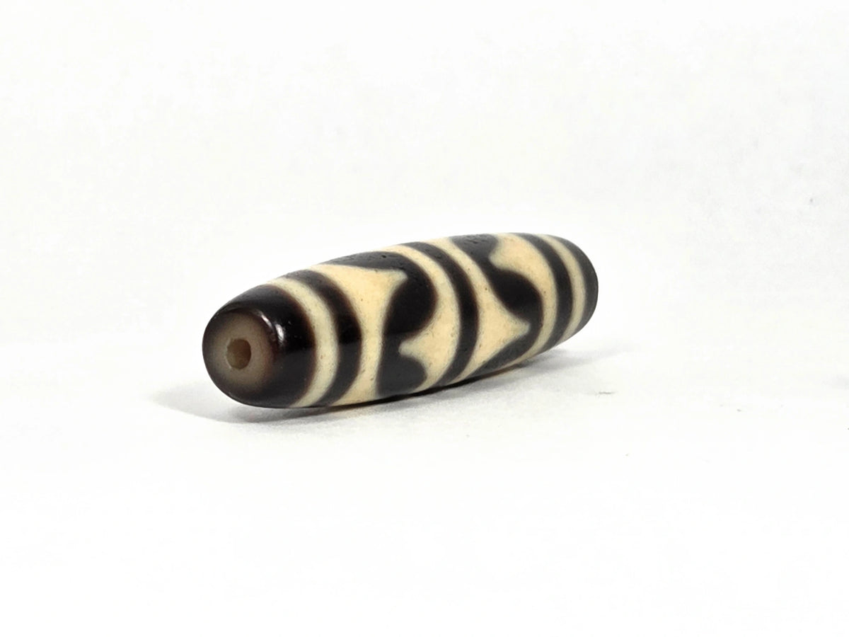 Opposite end view of antique Tibetan double wave dzi bead (AT-092826-DW2) showing second drill hole and warm brown end cap zone with black and cream wave pattern continuing to the tip