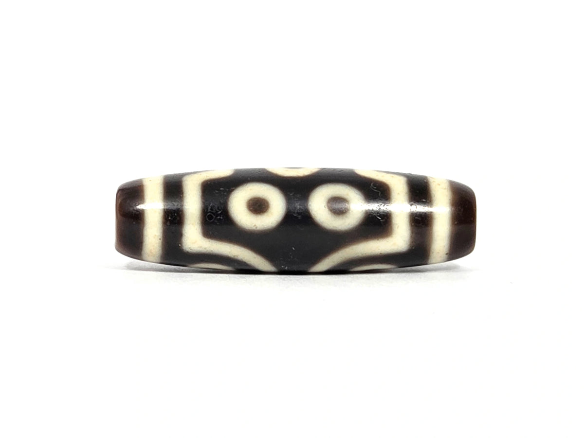Second lateral profile of antique seven-eye dzi bead showing two eyes flanked by angular bracket forms and maze-line connectors on dark brown-black ground, cream-white motif, 39.7 mm