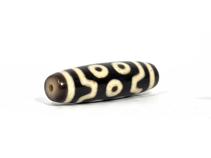 Opposite end view of antique Tibetan seven-eye dzi bead (AT-092826-7E) showing second drill hole and warm brown end cap zone with cream-white eye and wave motif continuing to the tip