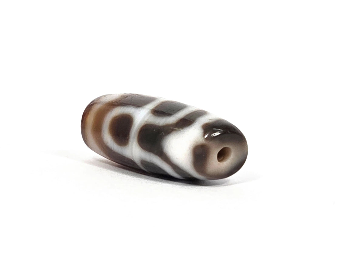 Angled end view of vintage Taiwan chalcedony dzi bead N-B-101426-TTL showing drill hole aperture at grey-toned chalcedony tip, cream motif bands visible close to tip, dark warm brown barrel body