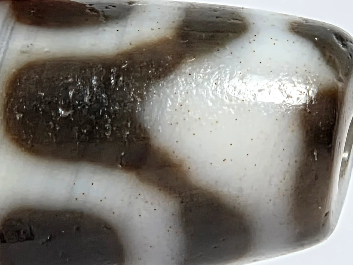 Macro close-up of vintage Taiwan chalcedony dzi bead N-B-101426-TTL showing bloodspot patina — fine red mineral speckling distributed across cream-white and dark zones of the motif surface; spots photograph as dark but are red in natural light